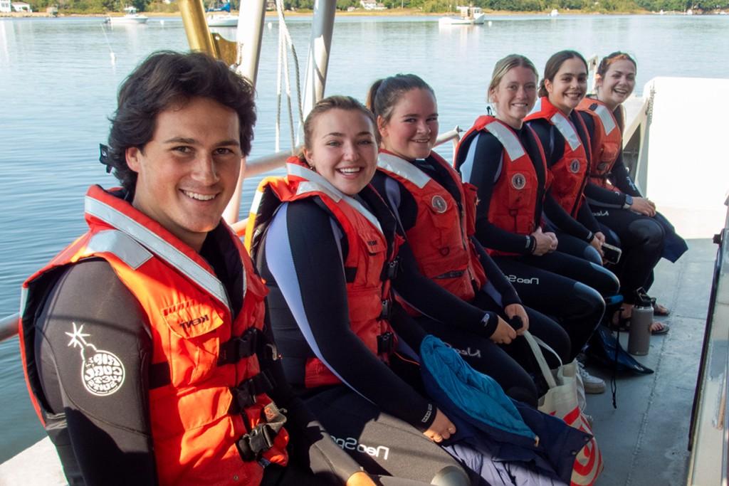 A group of students in orange life vests sit in a row on a U N E boat